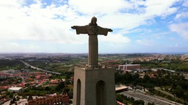 Portekiz 'in Almada kentindeki Cristo Rei heykeli kollarını açmış duruyor. Şehir manzarasında parçalı bulutlu bir gökyüzü altında binalar, yollar ve yeşil alanlar yer almaktadır..