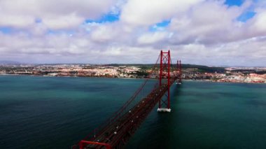 Lizbon, Portekiz 'deki 25 de Abril Köprüsü, Tagus Nehri üzerinde uzanır. Kırmızı asma köprü, derin mavi su ve şehir manzarasıyla çelişiyor..