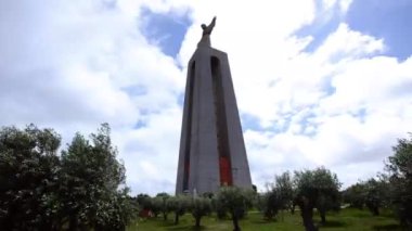 Portekiz 'in Almada kentindeki Cristo Rei heykeli ön planda zeytin ağaçları ile çevrili bulutlu bir gökyüzüne karşı açık kollarla ayakta duruyor..