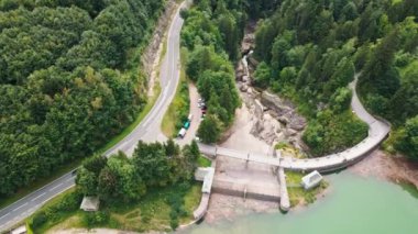Hava perspektifi, Avusturya 'da bir ormanın içinden geçen, doğanın ortasında altyapıyı vurgulayan küçük bir baraj ve park edilmiş araçlarla çevrili bir yol ortaya çıkarıyor..