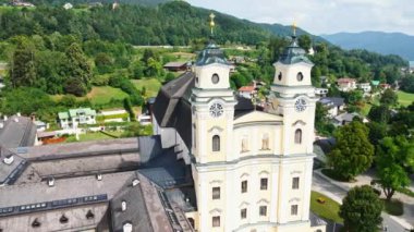 Mondsee Manastırı 'nın ikiz kuleleri kayan tepelere ve ormanlara karşı yükseliyor, konut binaları ve açık alanlar ön planda ve dağların ötesinde.