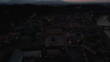 Salzburg, Avusturya 'ya ait bir hava manzarası, geceleri aydınlatılmış Salzburg Katedrali. Şehir manzarasında tarihi mimari ve soluk dağ çizgileri yer alıyor..