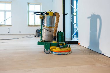 Zemin zımparalama makinesi, sarı ve yeşil gövdeli hafif ahşap bir zeminde duruyor. Odanın beyaz duvarları, geniş pencereleri ve görünür yapı iskelesi var..