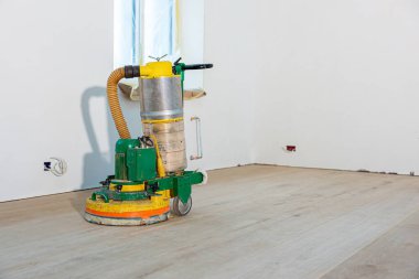 Toz torbasıyla yeşil ve sarı bir zımpara makinesi beyaz duvarlı ve küçük pencereli boş bir odada, açık ahşap bir zeminde duruyor..