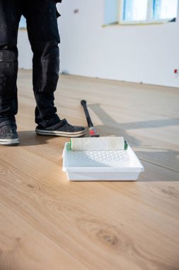 Ahşap bir zeminin yanında duran bir insan, tepside boya silindiri ile tedavi görüyor. Doğal ışık pencereden girer, tamamlanmamış duvarları vurgular..