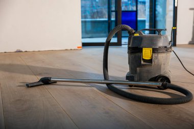 İçerideki ahşap zeminde sarı aksanlı ağır bir elektrikli süpürge oturuyor. Büyük cam kapılar, parmaklıkları ve bulanık yapıları olan bir açık hava sahası olduğunu gösteriyor..