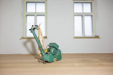 Beyaz duvarlı bir odada, açık ahşap bir zeminde, yeşil bir yer cilası makinesi. Koruyucu plastikli iki büyük pencere, sürekli bakım yapıldığını gösteriyor..