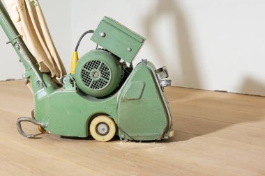 Kumaş bir toz torbasıyla eski yeşil yer cilalama makinesi açık tahta bir zeminde duruyor. Arka planda ince toz ve düz beyaz bir duvar görülebilir..