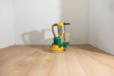 Beyaz duvarlı bir odada, açık ahşap bir zeminde yeşil ve sarı bir yer cilası makinesi duruyor. Parlak yüzey ışığı yansıtıyor, pürüzsüzlüğünü vurguluyor..