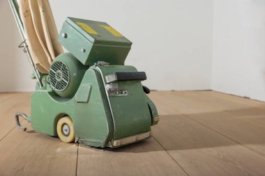 Metal gövdesi, lastik tekerlekleri ve bej hortumlarıyla yeşil zemin cilalama makinesi düz beyaz duvarlara karşı açık ahşap bir zeminde durur..