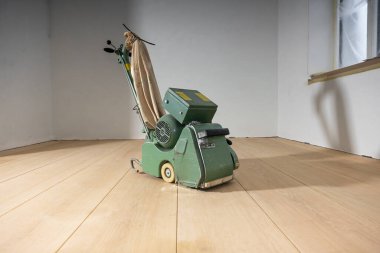 Yeşil zemin cilalama makinesi, beyaz duvarlı minimalist bir odada, açık ahşap bir zeminde ve kısmen bakım için kaplanmış bir pencerede bulunur..