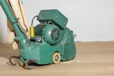 Yeşil bir yer cilası makinesi, bir motor, toz torbası ve ahşap bir zemine kablolar. Kısmen cilalanmış yüzey ve toz görünür..