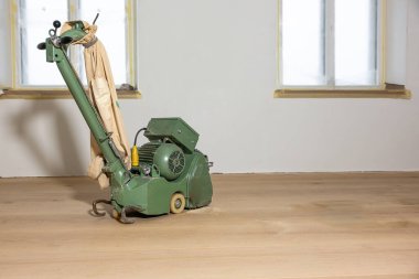 Pürüzsüz ahşap zeminde bej bir toz torbasıyla yeşil zemin cilası makinesi duruyor. İki bantlı pencere ve düz ışık duvarı, sürekli bakım yapıldığını gösteriyor..