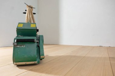 Elinde toz torbası olan yeşil bir yer cilası makinesi parlak, ahşap bir zeminde duruyor. Odanın düz beyaz duvarları var ve boş görünüyor..