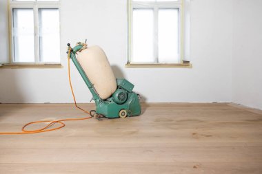 Bantlanmış camları ve turuncu elektrik kablosu olan beyaz duvarlı bir odada bej renkli bir toz torbasıyla yeşil bir parlatma makinesi..