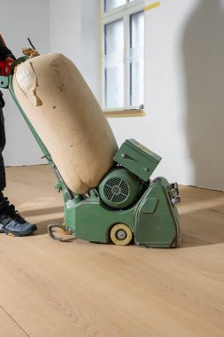 Bir işçi, beyaz duvarlı bir odada, açık ahşap bir zeminde yeşil bir parlatma makinesi kullanıyor. Bantlanmış pencere ve yenileme ayrıntıları görünür.