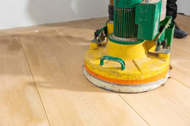 Sarı ve yeşil zemin cilalama makinesi, ışığı yansıtan, açık ahşap bir zeminde aktif olarak çalışır. Siyah ayakkabılı biri, beyaz bir duvarın yanında çalıştırıyor..