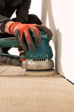Beyaz bir duvarın yakınındaki açık ahşap zeminde, ahşap dokusunu ve işin hassasiyetini vurgulayan yeşil bir elektrikli yer cilası kullanan bir insan..
