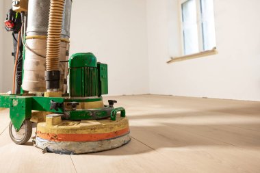 Yeşil ve sarı zemin parlatıcı bir makine, beyaz duvarlı bir odada, açık ahşap bir zeminde çalışır. Güneş ışığı tek bir pencereden akar..