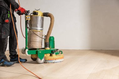 Siyah elbiseli bir işçi, açık ahşap bir yüzey üzerinde, görünür araçlar, kırmızı eldivenler ve mavi koruyucu ayakkabılar ile yer parlatıcı bir makine kullanır..