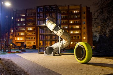 Nighttime view of a playground with a tall spiral slide, yellow play element, paved walkway, and a lit residential building in the background. clipart