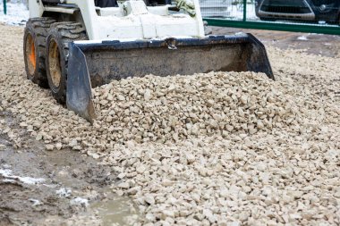 Küçük bir yükleyici çamurlu bir zemine hafif renkli çakıllar bırakıyor. Kirli kaplama tekerlekler ve yeşil metal çitler açık havada aktif bir çalışma alanı olduğunu gösteriyor..