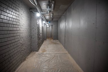 Bir tarafında açık tuğla, beton duvarlar, görünür borular ve kısmen açık bir kapı girişine açılan plastik kaplı bir zemin bulunan loş ışıklı koridor..