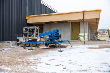 Kış boyunca bir inşaat sahasında mavi mobil bir asansör, kısmen inşa edilmiş siyah bir dış yüzey ve istiflenmiş yalıtım malzemesi bulunur..