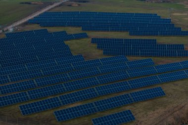 Açık bir alanda metal çerçeveler üzerinde sıralı koyu mavi paneller içeren büyük bir güneş çiftliği, arka planda görünen elektrik hatlarıyla çevrili çitlerle çevrili..