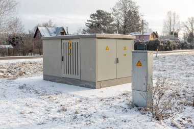 Karla kaplı bir alanda sarı uyarı işaretleri, bir kontrol paneli, seyrek bitki örtüsü ve arka planda eğimli çatıları olan evler bulunan bir hizmet kutusu yer almaktadır..