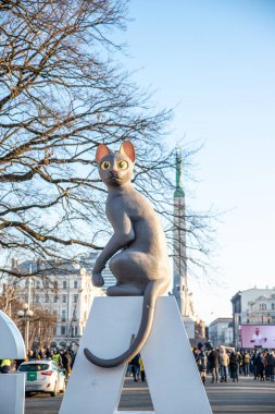 Letonya 'nın Riga kentindeki Özgürlük Anıtı' nın yakınındaki beyaz bir yapıda, çıplak ağaçlar ve kalabalık tarafından çerçevelenmiş büyük bir kedi heykeli bulunuyor..