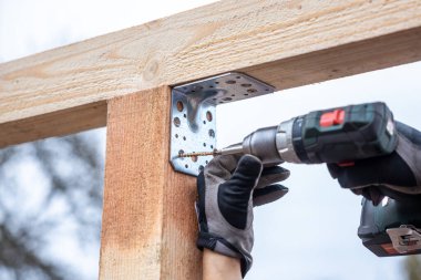 Bir insan, metal bir desteyi önceden delinmiş deliklerle ahşap bir kirişe tutturmak için matkap kullanır. Koruyucu eldivenler giyilir ve arka planda bir ağaç vardır..