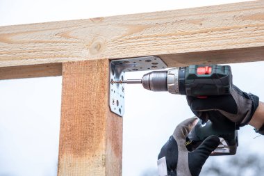 Bir insan, dışarı tahta kirişlere metal bir deste takmak için kablosuz matkap kullanır. Arka planda koruyucu eldivenler ve bulutlu bir gökyüzü görülüyor.