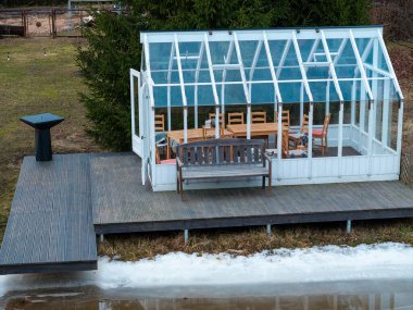 Camdan çatılı beyaz çerçeveli bir sera, çimlerle çevrili donmuş bir su kütlesinin yanında, ahşap bir platformda oturur..