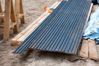 Açık hava inşaat sahasında çivili ahşap tahtalar, testere atlar, aletler, turuncu işaretler ve toprakla kaplı mavi bir muşamba var..