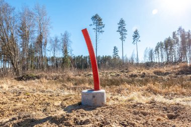 Bir inşaat sahasında, seyrek ağaçlar, kuru otlar ve açık mavi gökyüzü ile çevrili, ışık direği kurulumu için beton bir zeminden çıkan kırmızı bir kanal yer alır..