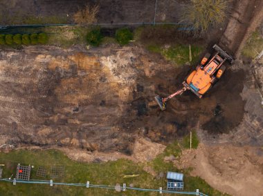 Hava görüntüsü, çimen ve seyrek bitki örtüsüyle çevrili, görünür çit ve malzemelerle çevrili bir inşaat sahasında çalışan turuncu bir kazıcı gösteriyor..
