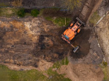 Bir turuncu kazıcı açık toprak, seyrek bitki örtüsü, küçük ağaçlar ve kısmen çimlerle kaplı görünen bir yerde çalışır..
