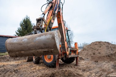 Çamurlu kovayla turuncu bir kazıcı toprak yığınları, lastik izleri, ağaçlar ve karanlık çatılı bir yapıyla çevrili bir inşaat sahasında oturur..
