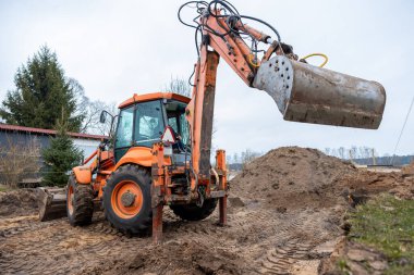 Bir turuncu kazıcı toprak inşaat alanında çalışır, kovası toprakla yükseltilir. Lastik izleri, toprak tepecikleri, kırmızı çatılı bir yapı ve ağaçlar görülebilir..