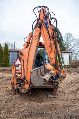 Görünür hidrolik hortumları olan turuncu bir kazıcı inşaat sahasında duruyor. Kırmızı çatılı beyaz bir bina ağaçlar ve çitlerle çevrilidir..