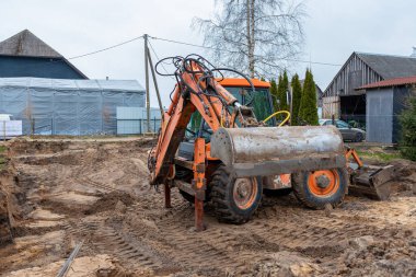 Turuncu bir kepçe doldurucu, arka planda lastik izleri olan, kırsal tarzda binalarla çevrili, yapıya benzer bir ahır ve elektrik direkleriyle dolu bir çamurda oturur..