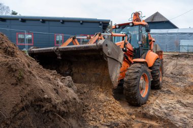 Turuncu bir kazıcı çamurlu zeminde, siyah duvarları, kırmızı çerçeveli pencereleri ve metal çitleri olan modern bir binanın yanında lastik izleri ile çalışır..