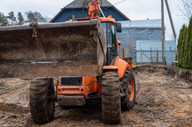 Çamurlu zeminde kaldırılmış kovası olan, lastik izleriyle çevrili turuncu bir kepçe doldurucusu. Arka planda mavi bir ev, metal çitler ve ağaçlar görünüyor..