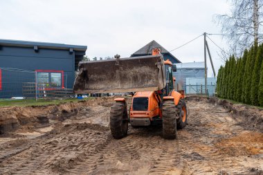 Ön yükleyici kovası olan turuncu bir inşaat aracı toprak bir alanda duruyor. Modern gri bir bina, yemyeşil ağaçlar ve elektrik direkleri görülebilir..