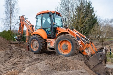 Turuncu bir kepçe doldurucusu arka kazma kolu genişletilmiş bir toprak tümsekte çalışır. Çıplak ağaçlar ve seyrek bitki örtüsü daha soğuk bir mevsim olduğunu gösteriyor..