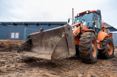 Elinde yıpranmış kova olan turuncu bir cephe doldurucusu toprak bir inşaat sahasında duruyor. Arka planda kırmızı pencereli ve çitli mavi bir bina var..