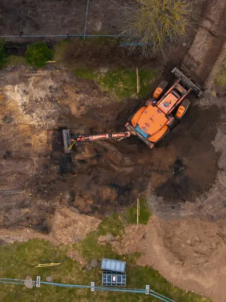 Hava perspektifi, çimenler, ahşap kazıklar, seyrek bitki örtüsü ve mavi borularla çevrili karanlık toprağı kazan bir turuncu kazıcıyı gösteriyor..