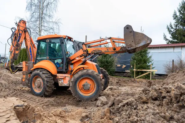 Turuncu bir kepçe yükleyici, kaldırılmış kova, bozulmuş toprak, lastik izleri, yemyeşil ağaçlar ve beyaz bir bina ile bir inşaat alanında çalışır..