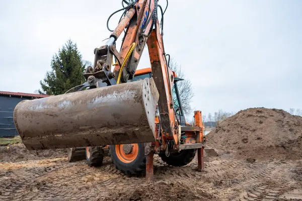 Çamurlu kovayla turuncu bir kazıcı toprak yığınları, lastik izleri, ağaçlar ve karanlık çatılı bir yapıyla çevrili bir inşaat sahasında oturur..